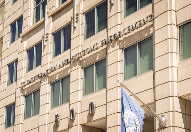 Facade of U.S. Immigration and Customs Enforcement building with agency letters and number 500, and a department flag flying in front.
