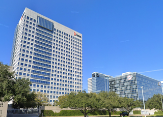Tall, modern office buildings with reflective glass windows under a clear blue sky, surrounded by green trees.