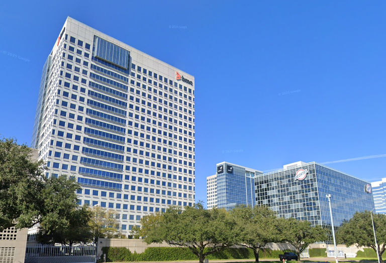 Tall, modern office buildings with reflective glass windows under a clear blue sky, surrounded by green trees.