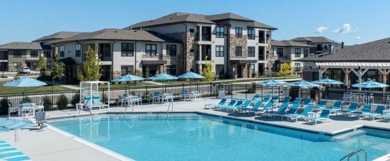 Outdoor pool area with blue lounge chairs and umbrellas in front of modern apartment buildings under a clear blue sky.