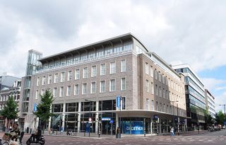 Modern brick and glass building on a city street corner, featuring large windows and a Decathlon store on the ground floor.