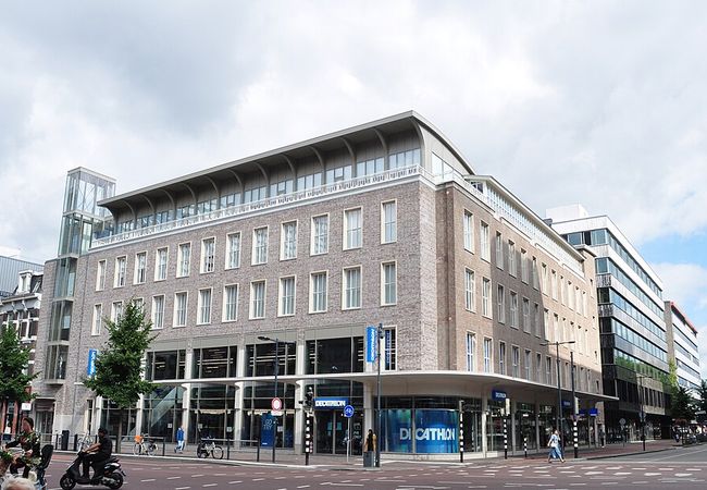 Modern brick and glass building on a city street corner, featuring large windows and a Decathlon store on the ground floor.