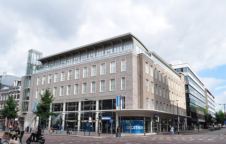 Modern brick and glass building on a city street corner, featuring large windows and a Decathlon store on the ground floor.