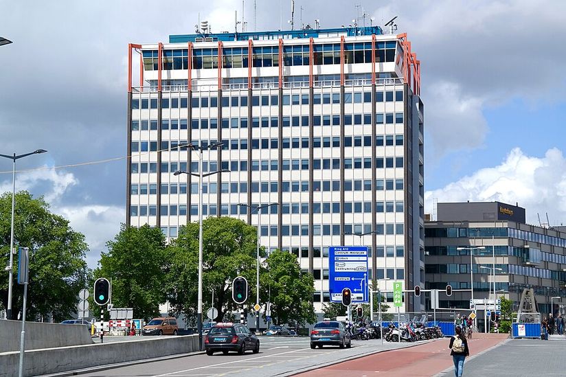 A tall office building with a grid of windows and orange accents, surrounded by trees and traffic lights, under a partly cloudy sky.