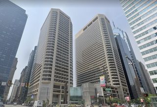 Tall twin buildings with numerous balconies in a busy urban setting, surrounded by other skyscrapers and street signs below.