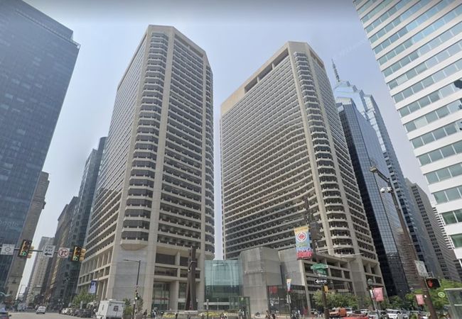 Tall twin buildings with numerous balconies in a busy urban setting, surrounded by other skyscrapers and street signs below.