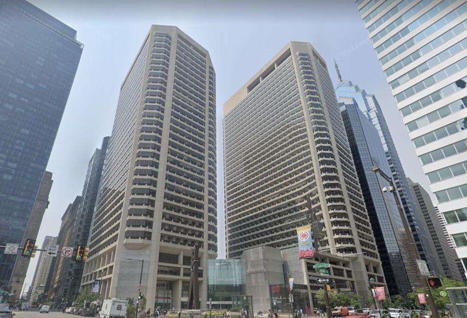 Tall twin buildings with numerous balconies in a busy urban setting, surrounded by other skyscrapers and street signs below.