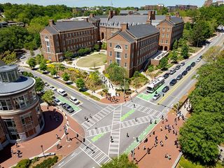 Aerial view of a busy urban intersection with distinctive brick buildings and green spaces, surrounded by cars and pedestrians on a sunny day.