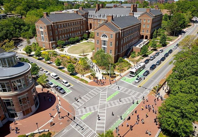 Aerial view of a busy urban intersection with distinctive brick buildings and green spaces, surrounded by cars and pedestrians on a sunny day.