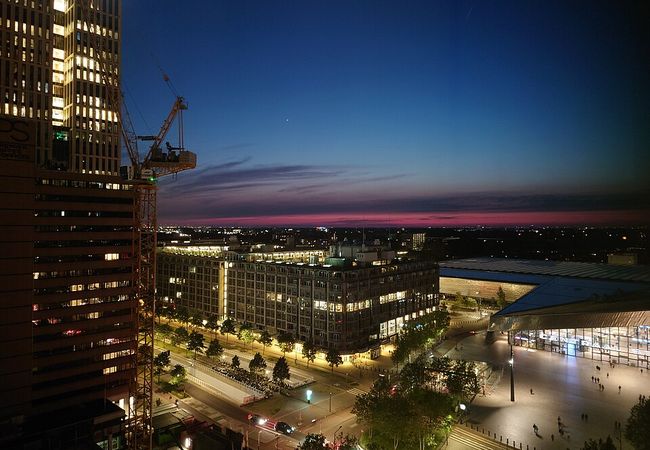 Cityscape at dusk with illuminated buildings, a construction crane, and a pink horizon beneath a deep blue sky.