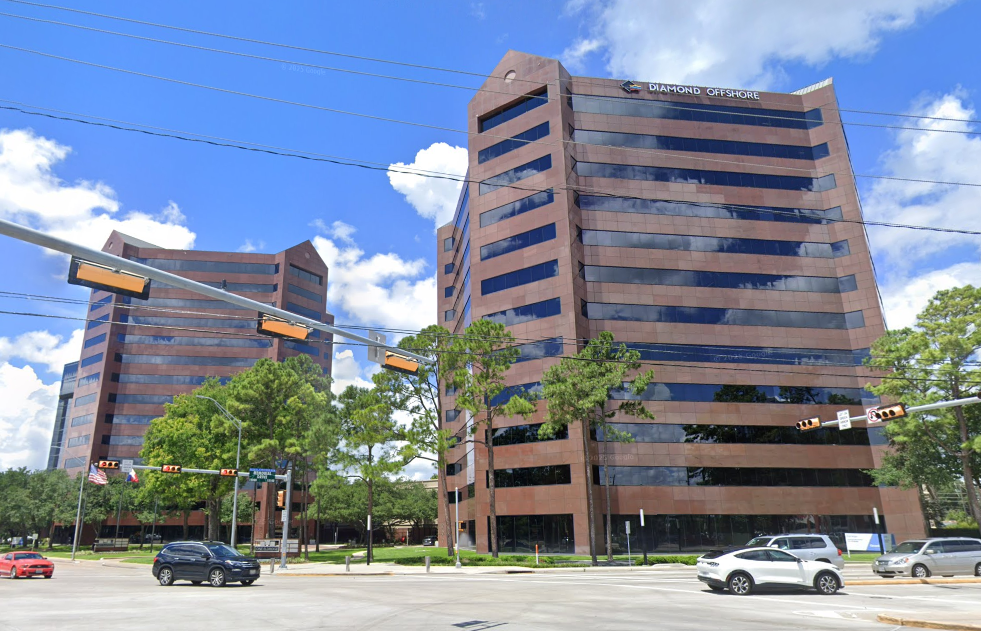 Two modern office buildings with reflective glass windows under a bright blue sky and white clouds, surrounded by trees and a few cars on the road.