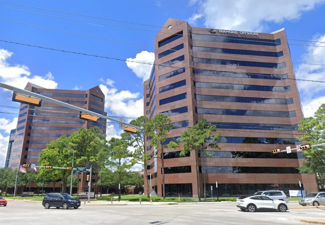 Two modern office buildings with reflective glass windows under a bright blue sky and white clouds, surrounded by trees and a few cars on the road.