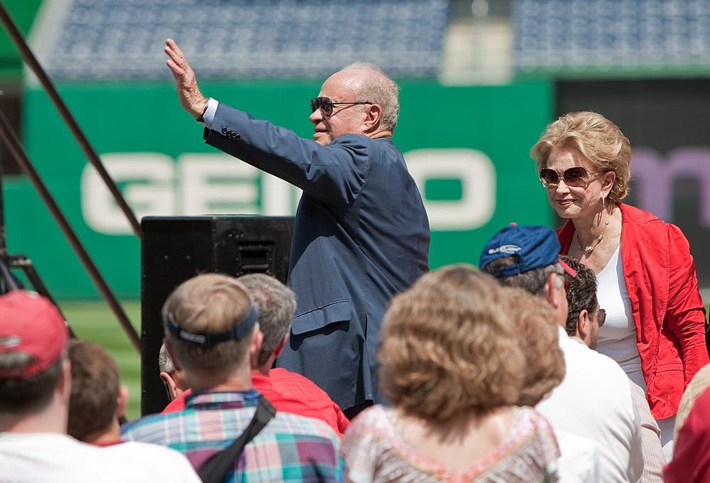 Ted Lerner in suit stands and waves in a baseball stadium with his wife, Annette, standing nearby