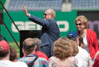 Ted Lerner in suit stands and waves in a baseball stadium with his wife, Annette, standing nearby