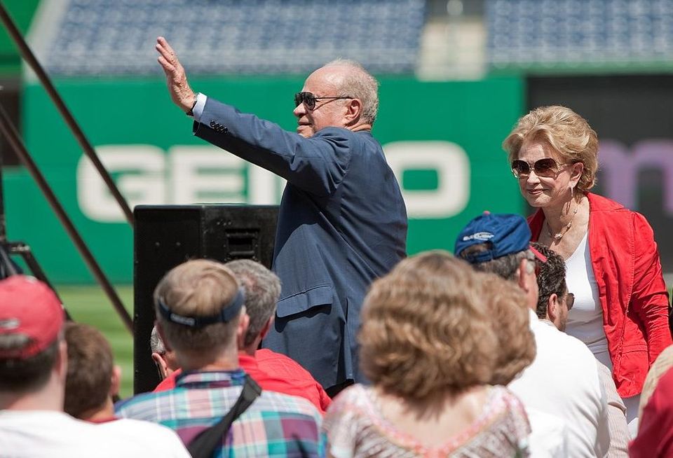 Ted Lerner in suit stands and waves in a baseball stadium with his wife, Annette, standing nearby