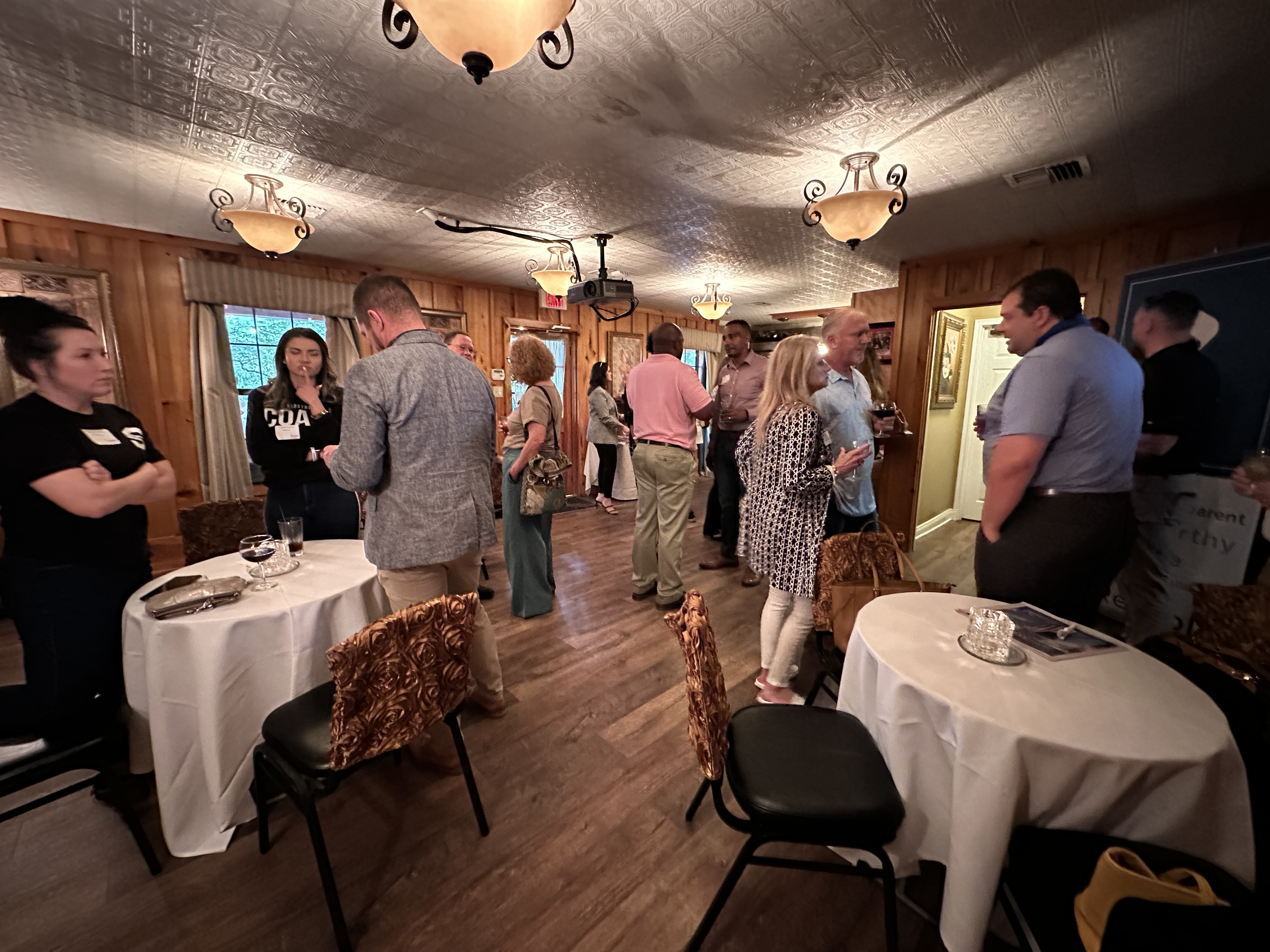 People mingling and conversing in a warmly lit room with wooden walls, tables, and chairs.