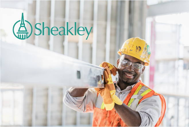 Photo of a man wearing a safety vest, a hard hat and safety goggles holding a metal beam and smiling. The photo has Sheakley's logo in the corner.