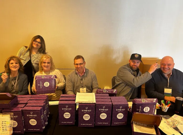 A group of six people smiling behind a table filled with stacked purple "Kynd Kit" boxes labeled "Financial Literacy."