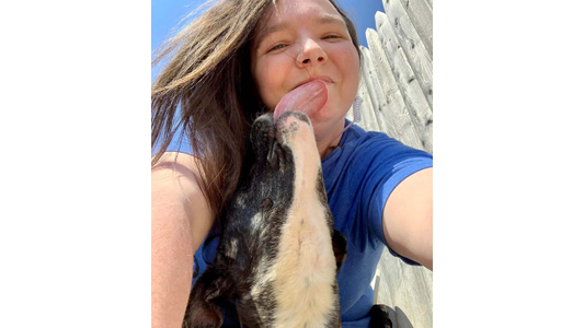 K9 Kaos Head Trainer Ashe Preston smiles as a black and white dog licks her face, set against a sunny blue sky and wooden fence background.