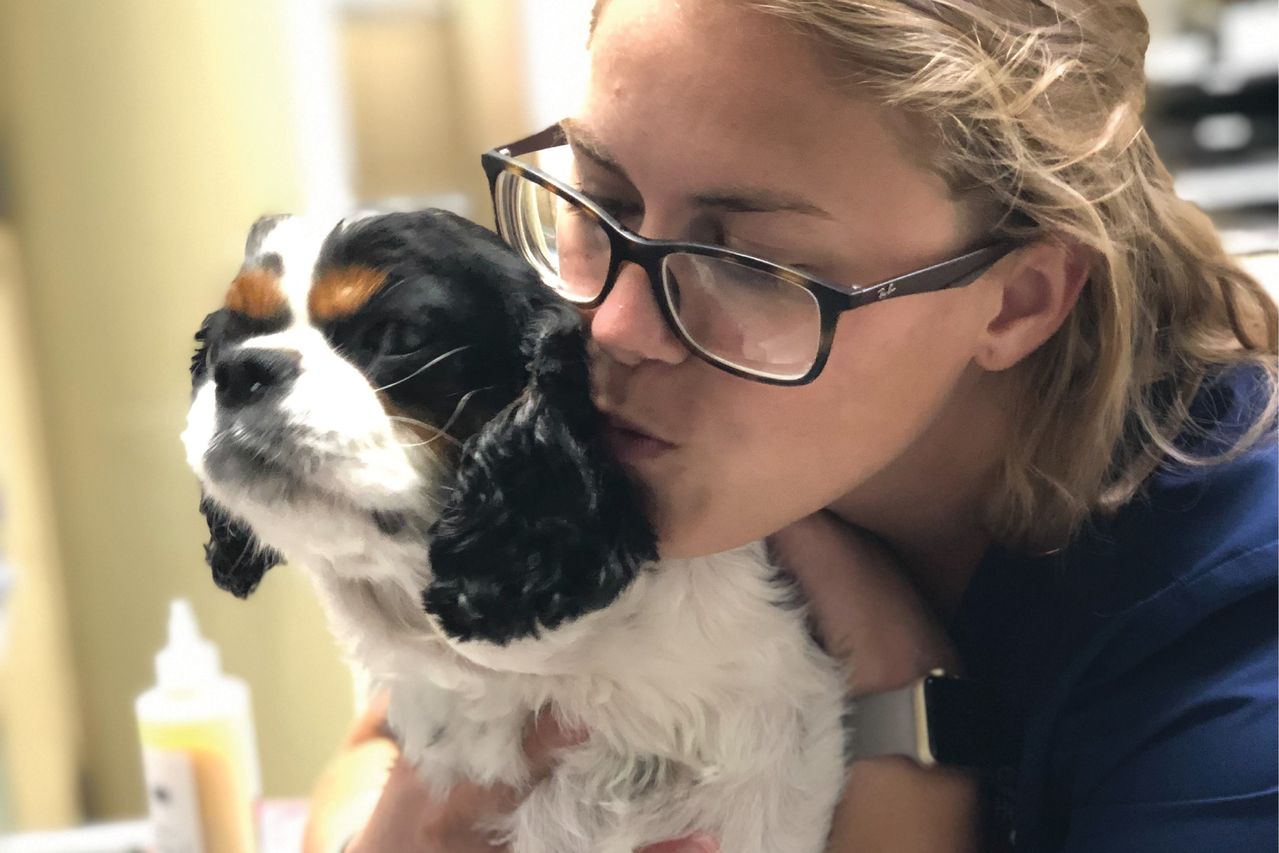 A female staff member wearing glasses hugs and kisses a small black and white dog with closed eyes, expressing affection and care.