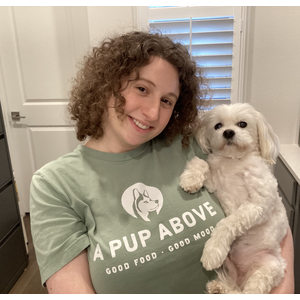 At Bark and Paw Groomer Ariana holding a small white dog in a kitchen. Wearing a green shirt with "A Pup Above" text and dog logo.