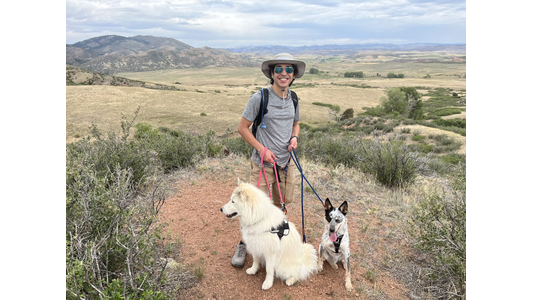 Dr. Zachary Subealdea standing on a scenic trail with two dogs, a white one and a spotted one, with mountains in the background