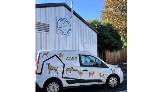 White van with dog illustrations parked outside a building labeled "Bow Wow Bungalow," featuring dog boarding and daycare service logo.