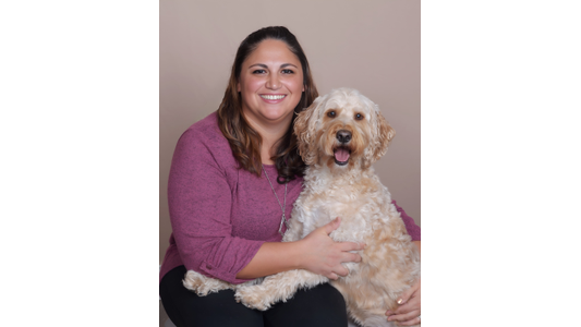 Dr. Nikki Medina - Medical Director smiles while hugging a fluffy, light-colored Labradoodle against a neutral background.
