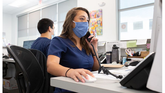 Vet team member wearing blue medical mask and scrubs talks on the phone while using a computer in an office setting. Another person works in the background.