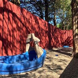 Blue plastic playground slide in a gravel area, bordered by a tall red wooden fence and surrounded by trees casting shadows.