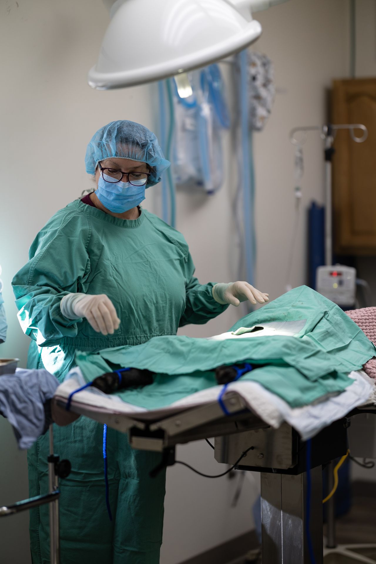 A surgeon in green scrubs and a mask stands next to an operating table in a brightly lit room, preparing for surgery.