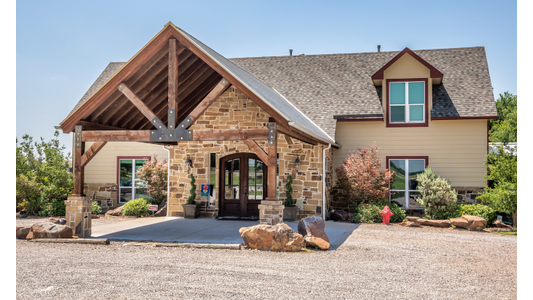 Rover Resort with a stone facade, wooden beams, and a covered entrance, surrounded by shrubs and small trees on a gravel driveway.