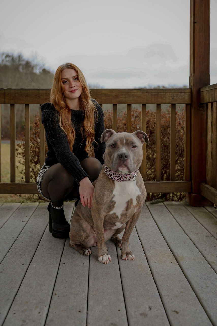 Gwen -Groomer kneels beside a large, gray and white pitbull on a wooden porch, with bushes and trees in the background.