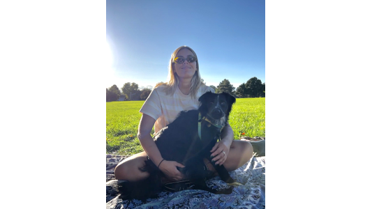 A woman wearing sunglasses sits on a blanket in a sunny park, holding a black dog on her lap, with green grass and trees in the background.