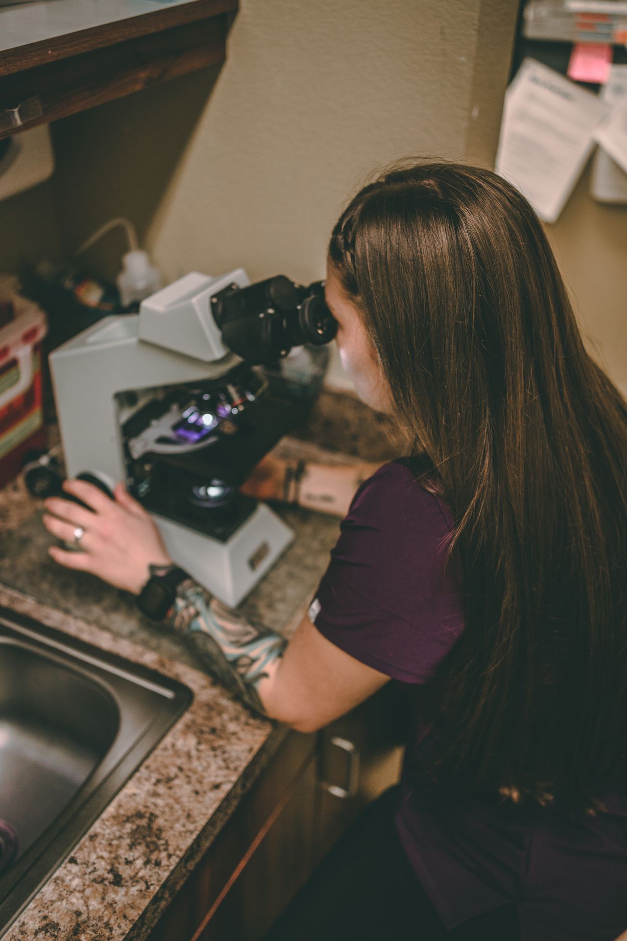 A woman with long brown hair examines a sample under a microscope on a kitchen counter, concentrating intensely.