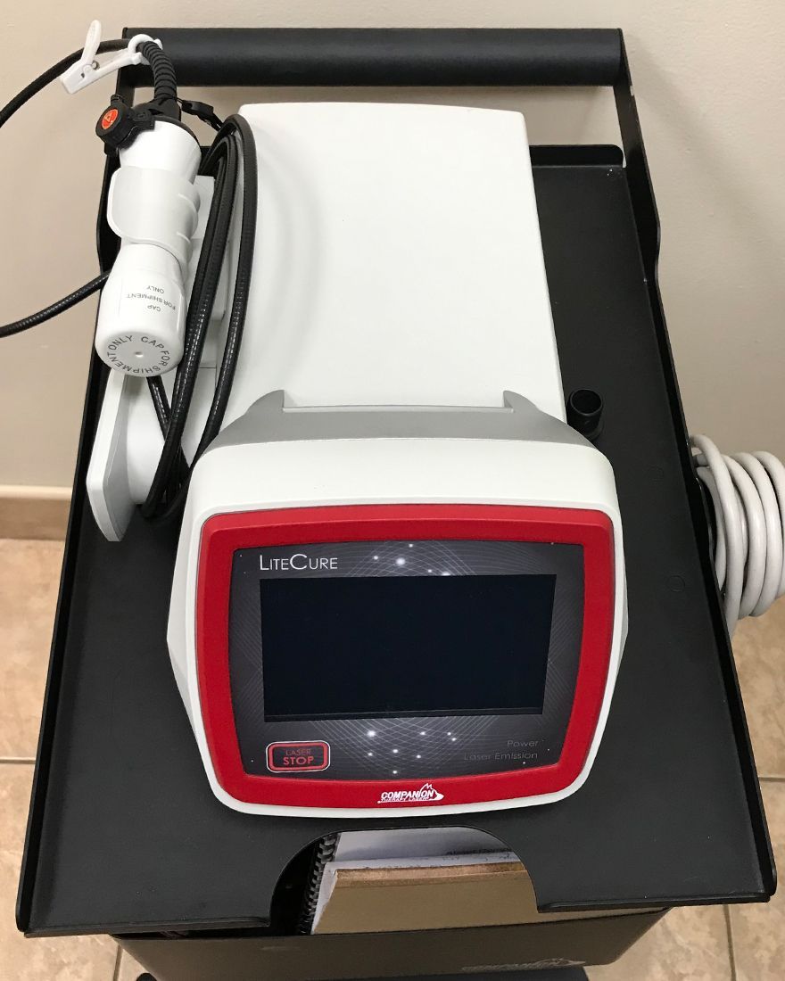 A LiteCure laser therapy device with a red and gray front panel, LCD screen, and white wand, placed on a black cart.