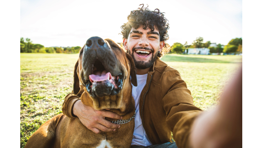 A joyful person takes a selfie with a large brown dog in a sunny park, both smiling widely.