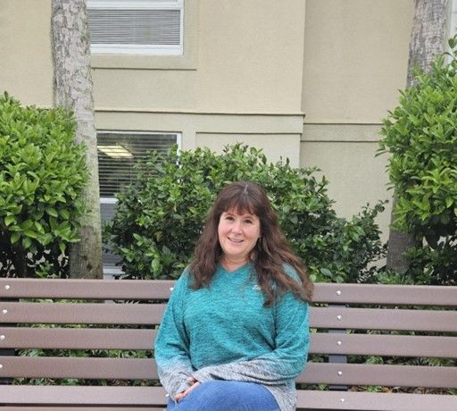 Lex -ACA sits on a bench in front of a beige building with green bushes.