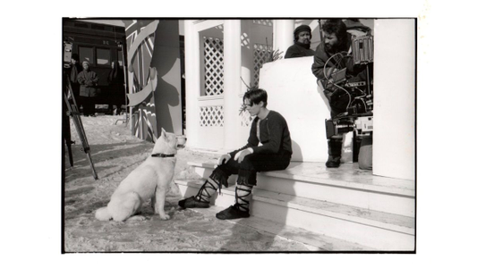 A man sitting on steps, looking at a white dog. A film crew with a camera is beside him. Snow covers the ground.