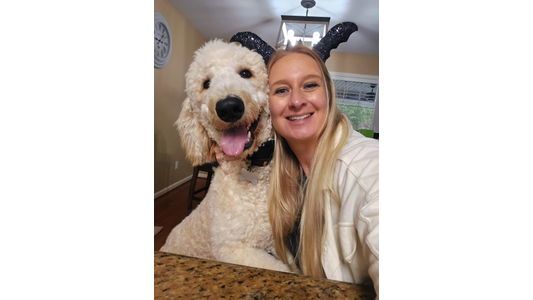 Greymont Kennel Jessica -District Director smiling beside a happy, fluffy dog wearing sparkly bat wings, sitting at a table in a warmly lit room.