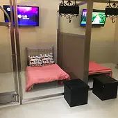 Grand Luxury suite with a single bed, pink bedding, decorative pillows, two black ottomans, and two wall-mounted TVs displaying different images.