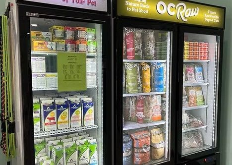 Commercial glass-door freezers in a pet store stocked with frozen raw dog and cat food, featuring brands like Answers Pet Food and OC Raw Dog