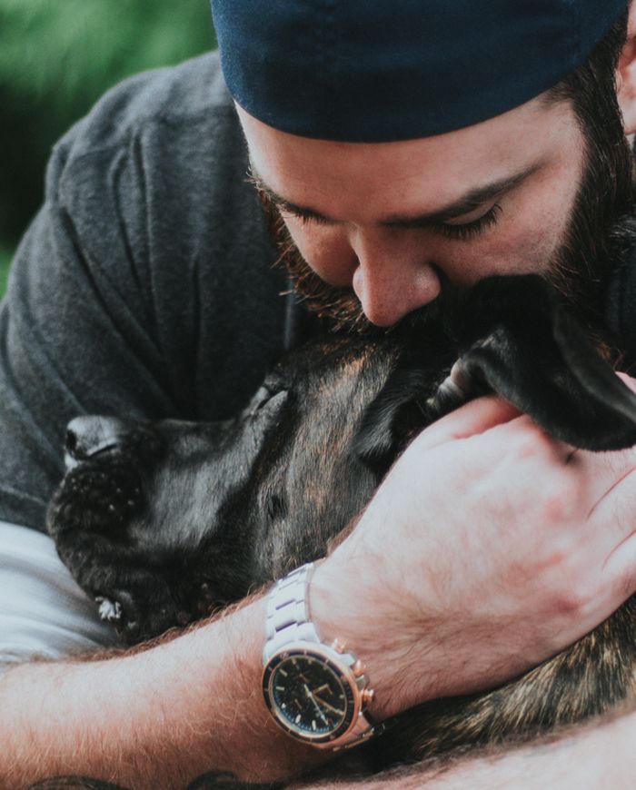 A man wearing a blue cap lovingly embraces a large, dark-coated dog, resting his head against the dog's, in a green outdoor setting.