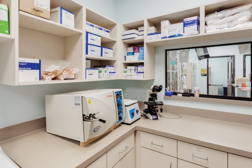 A laboratory with shelves full of medical supplies, an autoclave, and a microscope on a countertop, with a glass window showing another room.