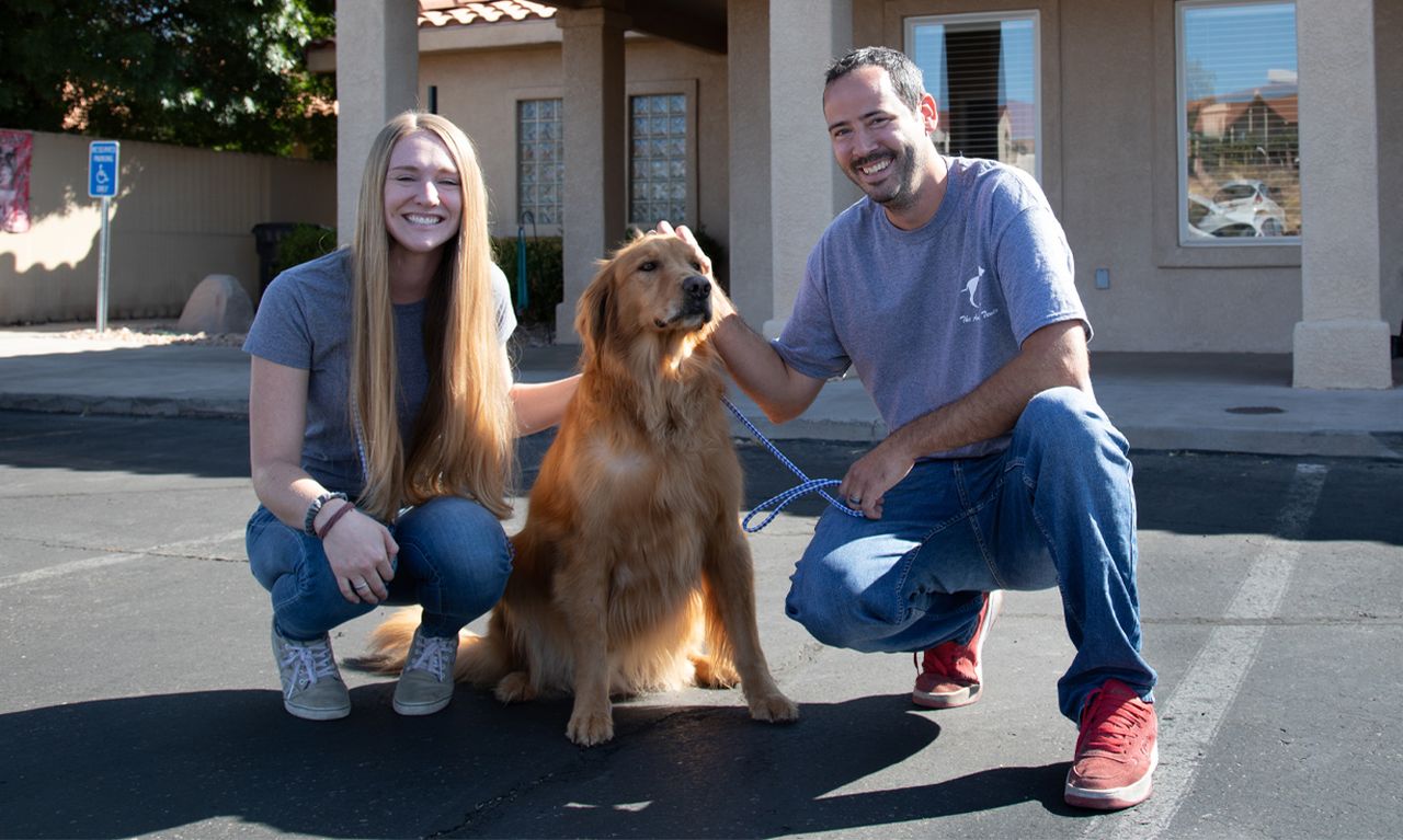 Animal Tender staff smiling and kneeling beside a golden retriever on a sunlit pavement in front of a building.