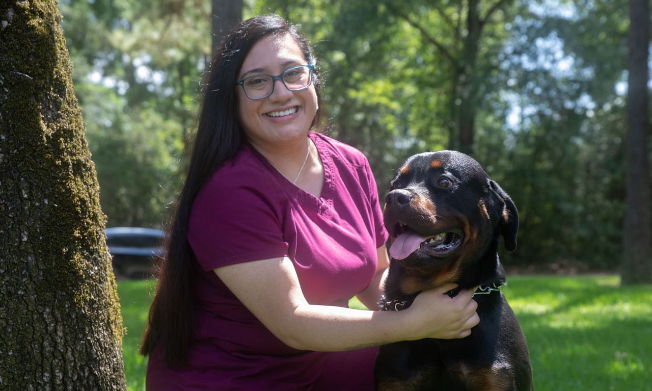 Kings Crossing staff member smiles while kneeling beside a Rottweiler in a lush, sunlit park.