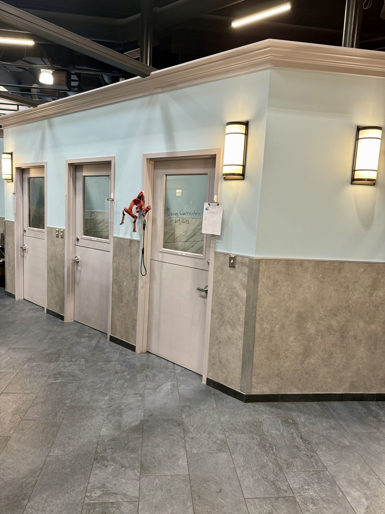 Interior dog kennels, light blue walls, beige lower paneling, and wall lamps. A stuffed animal hangs on the middle door.