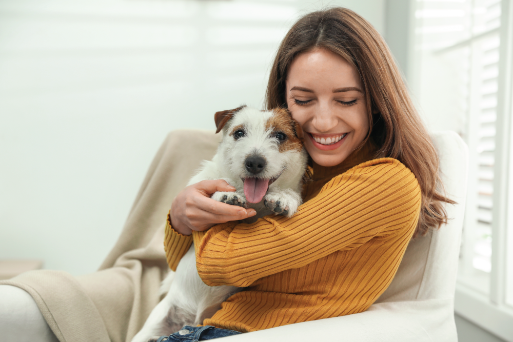 A smiling female staff member wearing a yellow ribbed sweater affectionately hugging a happy white and brown dog while sitting in an armchair indoors