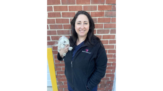 Brittany France -Assistant Manager & Veterinary Assistant holding a small white dog, standing in front of a brick wall, smiling at the camera.
