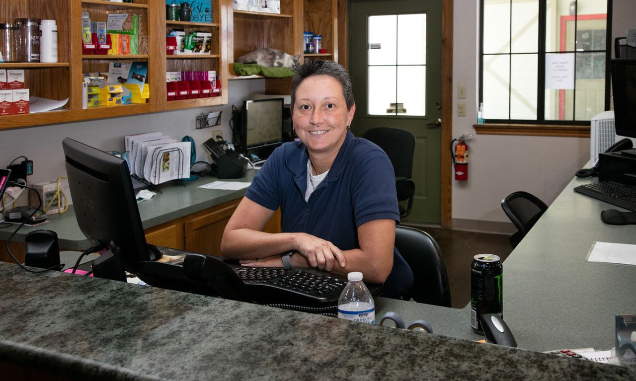 Hope Animal Clinic staff member smiling and sitting at a desk in an office, surrounded by various office supplies and shelves with products.